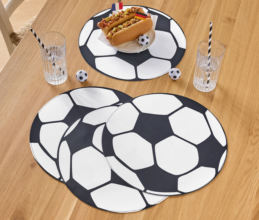Deux hot-dogs avec des drapeaux sur une assiette à motif de ballon de football, à côté de verres avec des pailles et une décoration de football sur une table en bois.