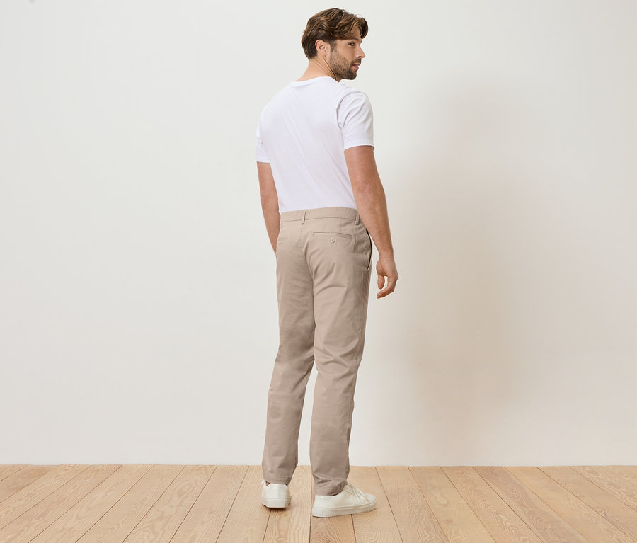Homme vu de dos portant un pantalon beige et un t-shirt blanc, debout sur un plancher en bois devant un mur blanc.