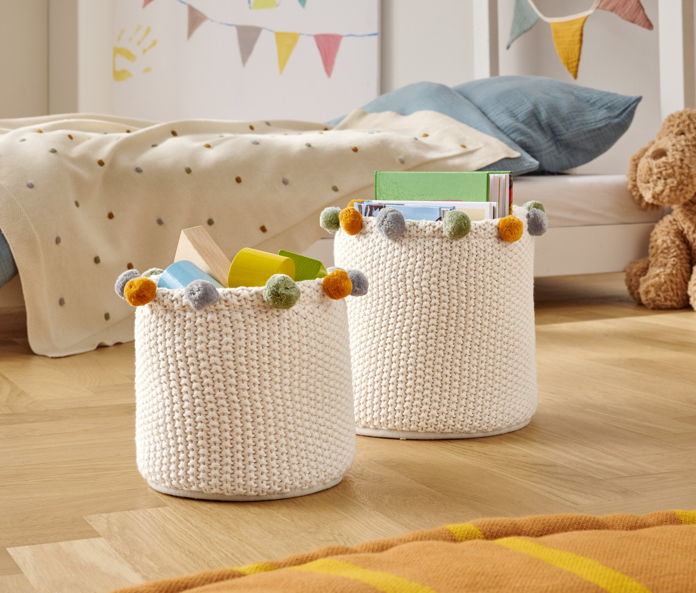 Deux paniers de rangement tricotés avec des pompons colorés sont posés sur un plancher en bois dans une chambre d'enfant.