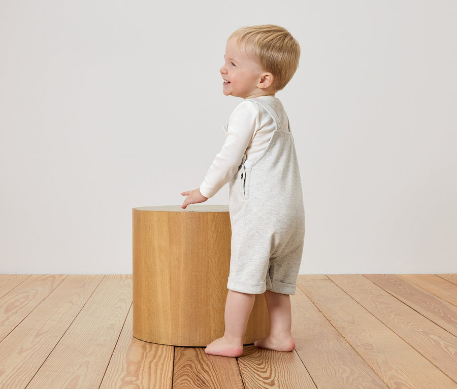 Un petit garçon en salopette gris clair se tient à côté d'un tabouret rond en bois.