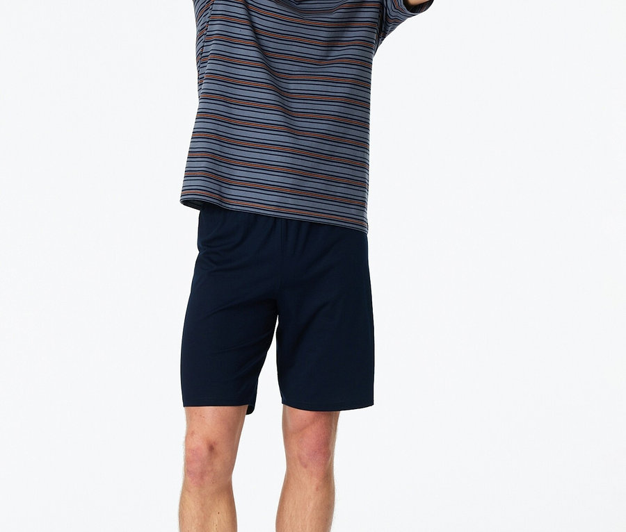 Un homme se tient pieds nus, les mains levées, vêtu d'un t-shirt rayé et d'un short bleu sur un fond blanc.