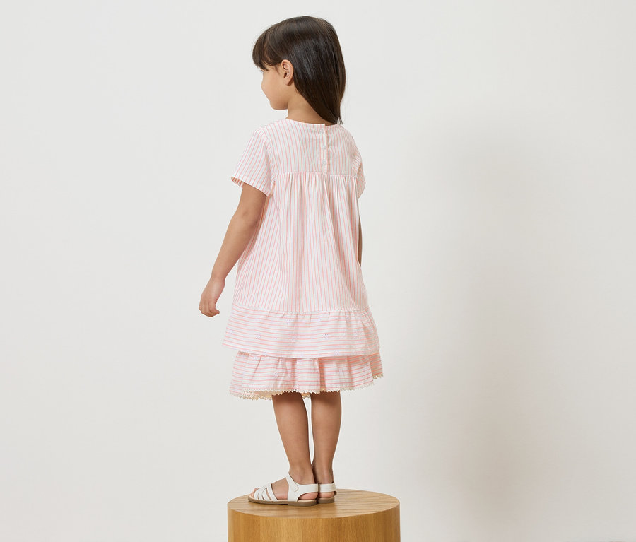 Fillette debout sur un tabouret, portant une robe rayée rose et blanche et des sandales blanches.