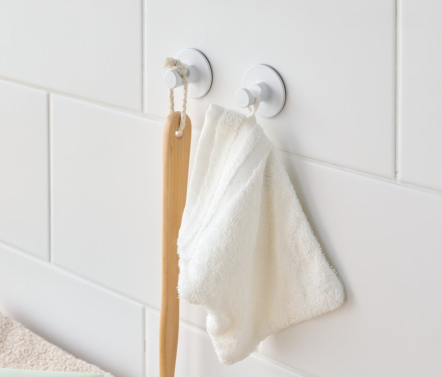 Une brosse de bain et une serviette blanche sont suspendues à des crochets blancs sur un mur carrelé blanc.