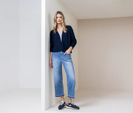 Une femme est appuyée contre un mur, portant un cardigan bleu marine, un jean bleu et des chaussures bleu marine.