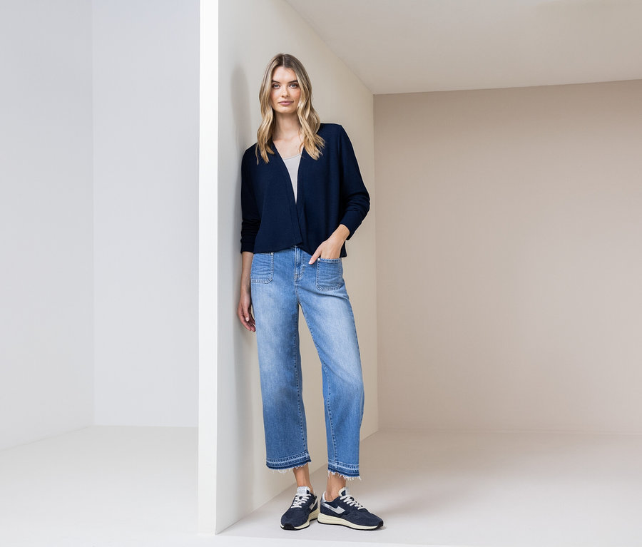 Une femme est appuyée contre un mur, portant un cardigan bleu marine, un jean bleu et des chaussures bleu marine.