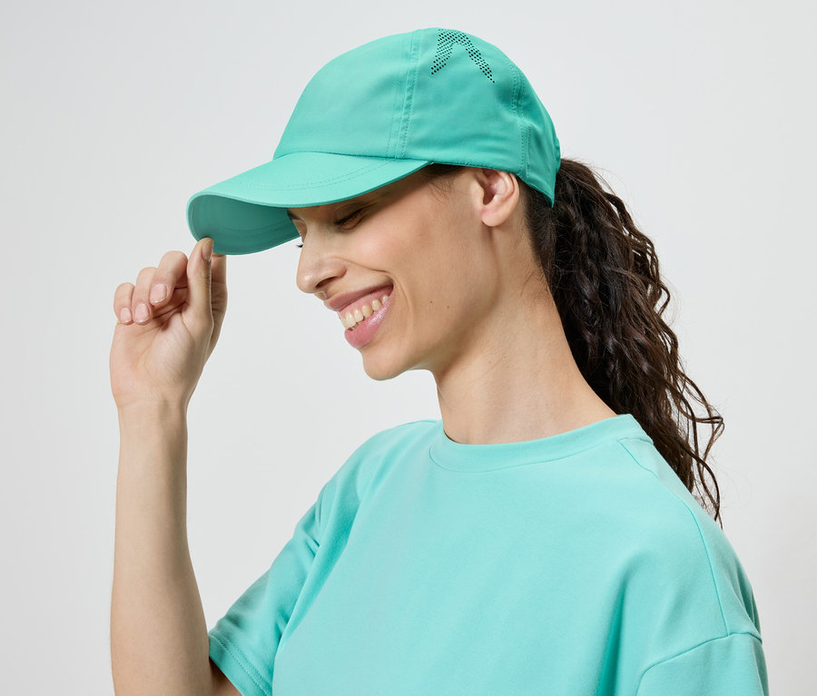 Une femme souriante portant une casquette et un t-shirt turquoise touche la visière de sa casquette.