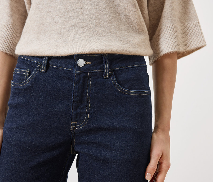 Gros plan d'une femme portant un jean foncé et un pull beige sur fond blanc.