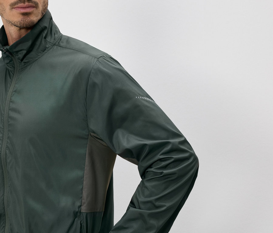 Homme portant une veste vert olive zippée à col montant, avec des empiècements latéraux en maille et des détails réfléchissants sur la manche.