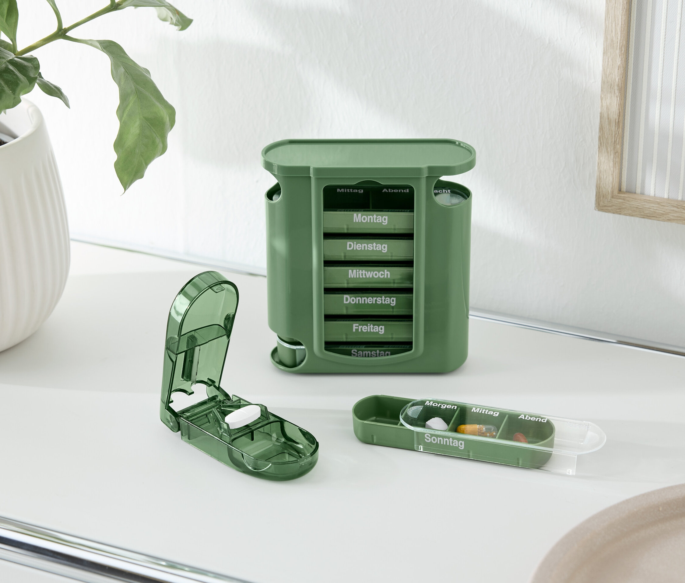 Sur une table, un pilulier vert, un coupe-comprimés et une boîte à pilules sont posés à côté d'une plante en pot.