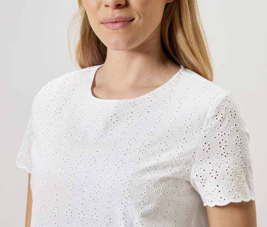 Une femme porte un chemisier blanc à broderie anglaise à manches courtes.