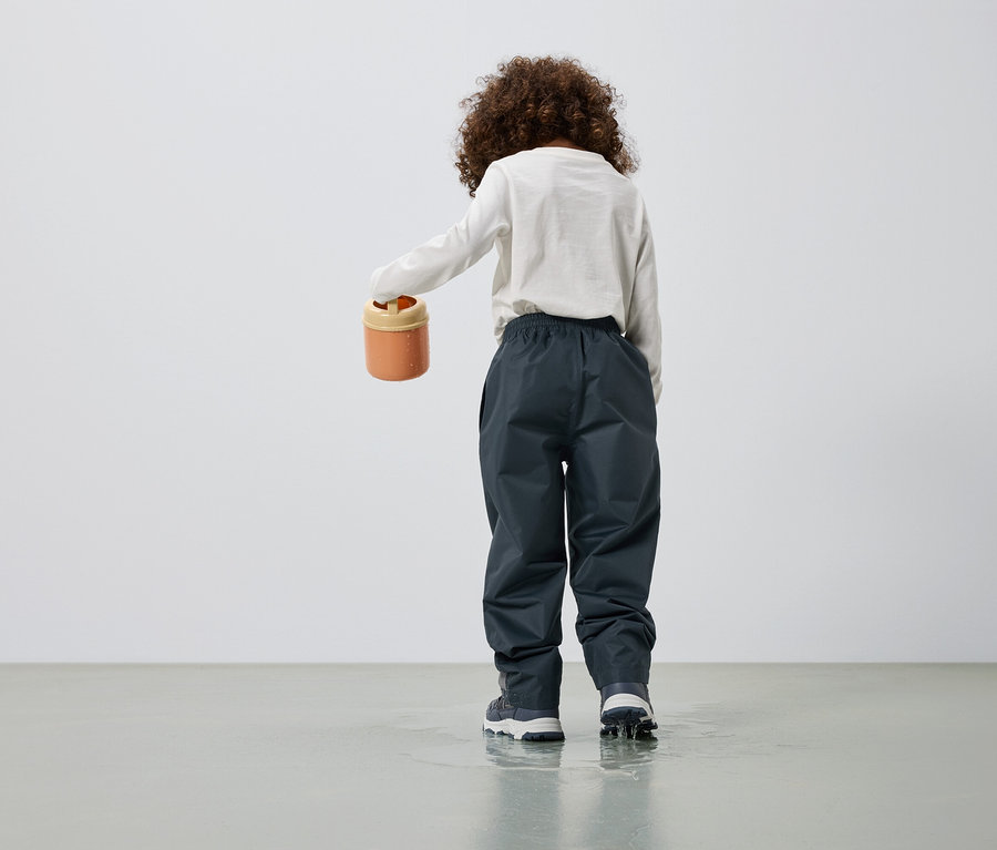 Enfant vu de dos tenant un seau orange, portant un pantalon anthracite et debout dans une flaque d'eau.