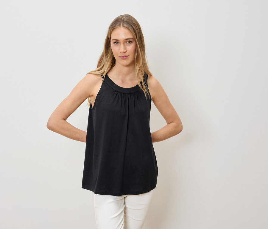 Une femme aux longs cheveux blonds porte un haut noir sans manches et un pantalon blanc, et pose les deux mains sur les hanches.