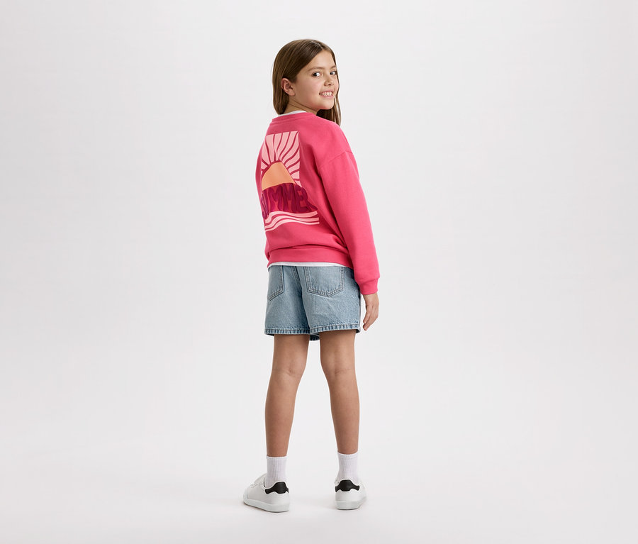 Fillette vue de dos, portant un sweat-shirt rose avec un motif de lever de soleil, un short en jean bleu et des baskets blanches.