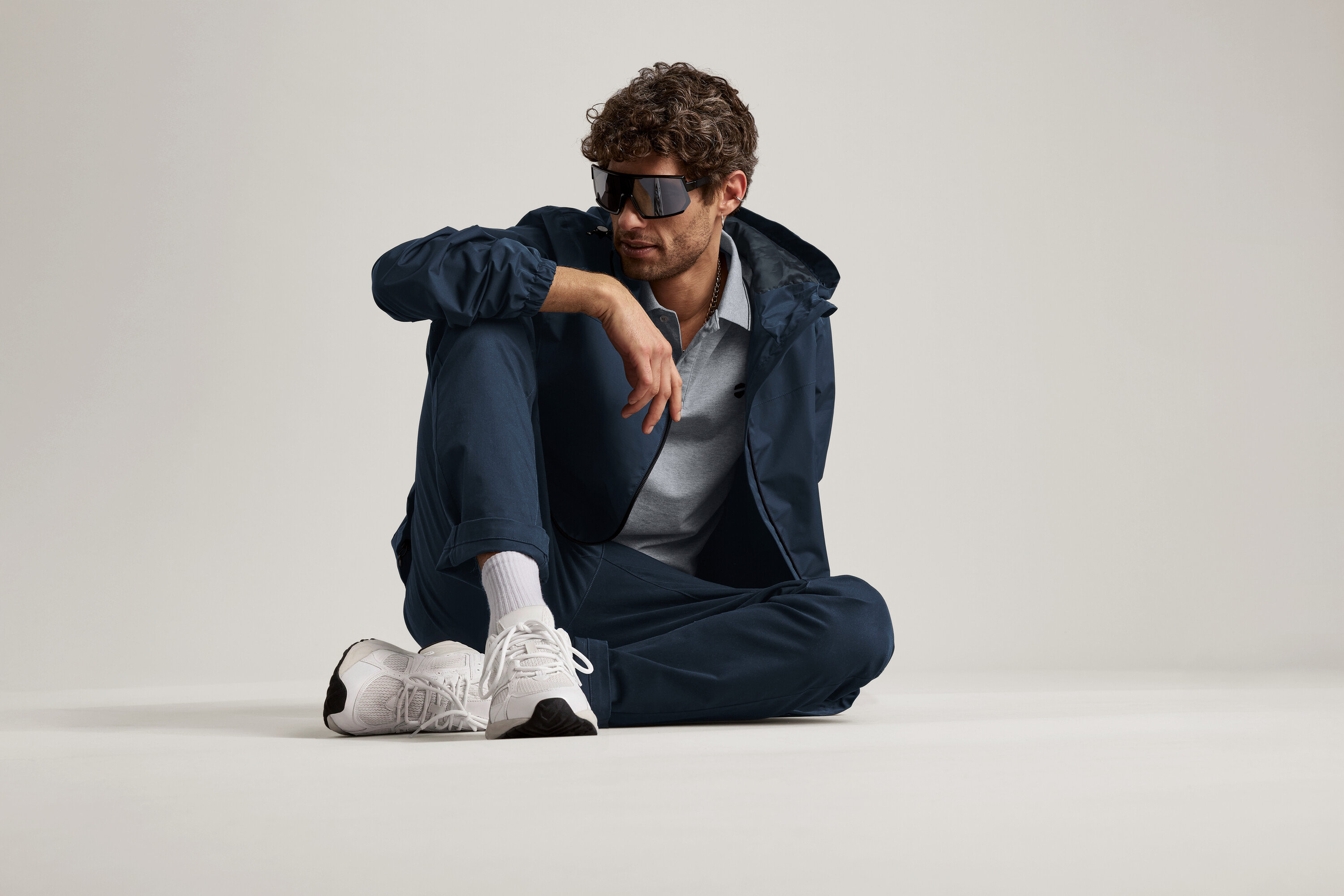 Un homme est assis par terre, portant un chino bleu marine, une veste et des lunettes de soleil.