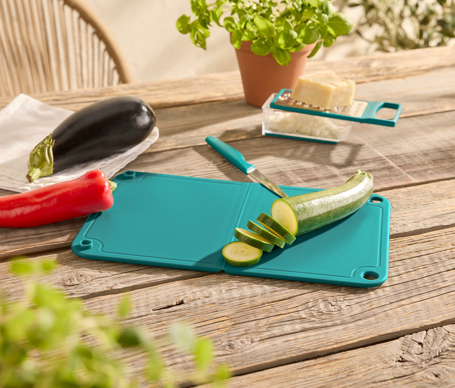 Sur une table en bois se trouvent un poivron rouge, une aubergine, une planche à découper verte avec un couteau et une courgette coupée, une râpe à fromage avec du fromage et un pot avec une plante.