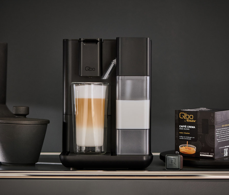 Un Qbo MILK MASTER Velvet Black prépare un latte macchiato. À côté se trouvent des capsules de café et une tasse.