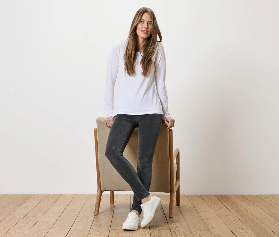 Une femme aux cheveux longs, portant un haut blanc, un pantalon gris et des chaussures blanches, s'appuie sur une chaise devant un mur blanc.