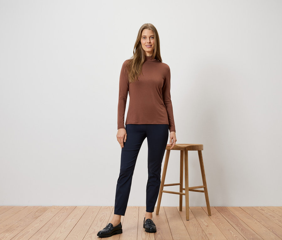 Une femme portant un pull à col roulé marron et un pantalon bleu pose à côté d'un tabouret sur un plancher en bois.