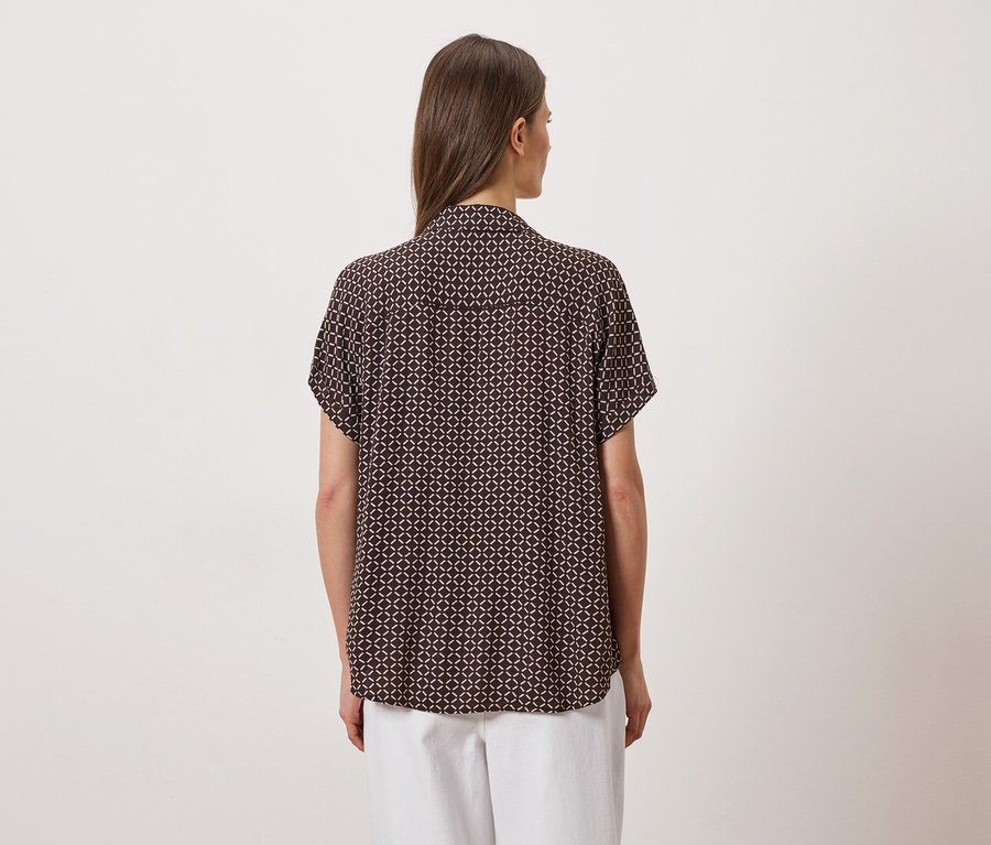 Vue arrière d'une femme aux cheveux bruns portant un chemisier marron à manches courtes avec un motif géométrique et un pantalon blanc.