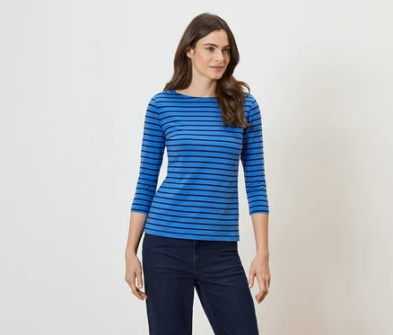 Une femme aux longs cheveux bruns ondulés porte un haut bleu à rayures horizontales et un jean foncé.