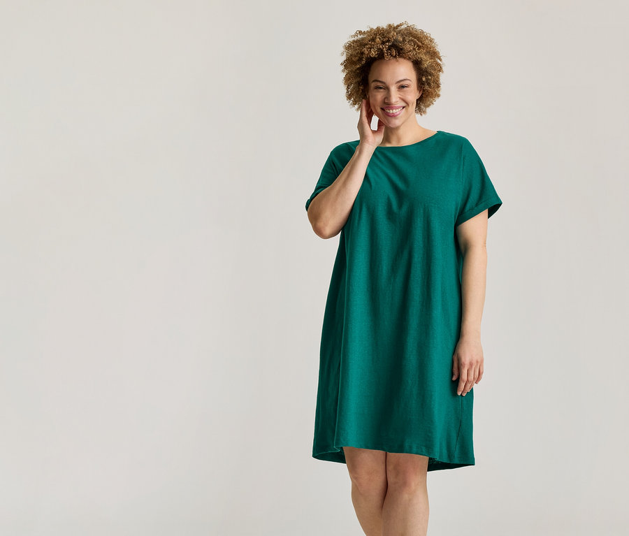 Femme souriante aux cheveux bouclés, vêtue d'une robe verte à manches courtes, touchant son visage.