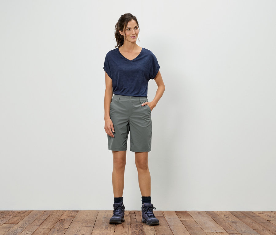 Une femme se tient devant un mur blanc, portant un t-shirt bleu, un short gris et des chaussures de randonnée.