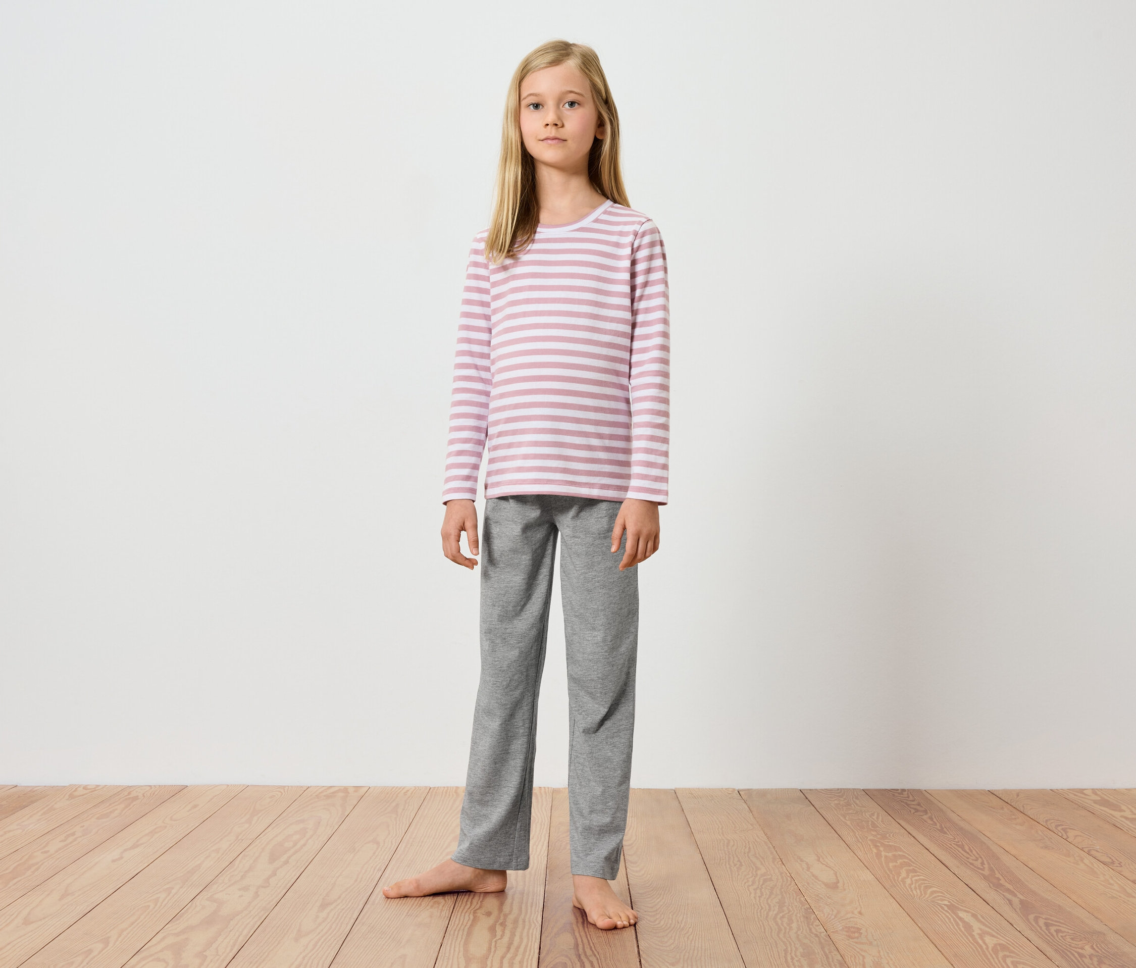 Une jeune fille se tient pieds nus sur un plancher en bois devant un mur blanc. Elle porte un haut à manches longues rayé rose et blanc et un pantalon gris.