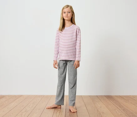 Une jeune fille se tient pieds nus sur un plancher en bois devant un mur blanc. Elle porte un haut à manches longues rayé rose et blanc et un pantalon gris.