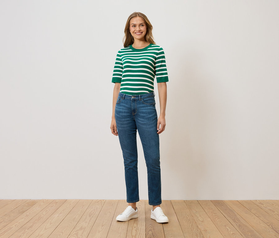 Une femme debout porte un haut rayé vert et blanc, un jean bleu et des baskets blanches.