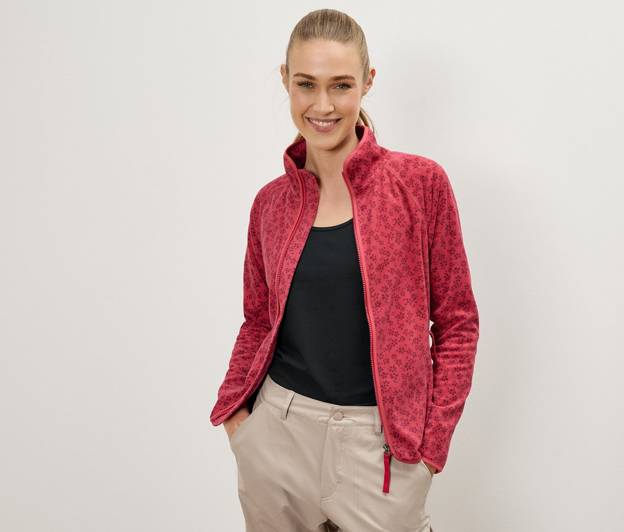 Une femme souriante pose devant un mur blanc, vêtue d'une veste polaire rouge à motifs floraux, d'un haut noir et d'un pantalon beige.