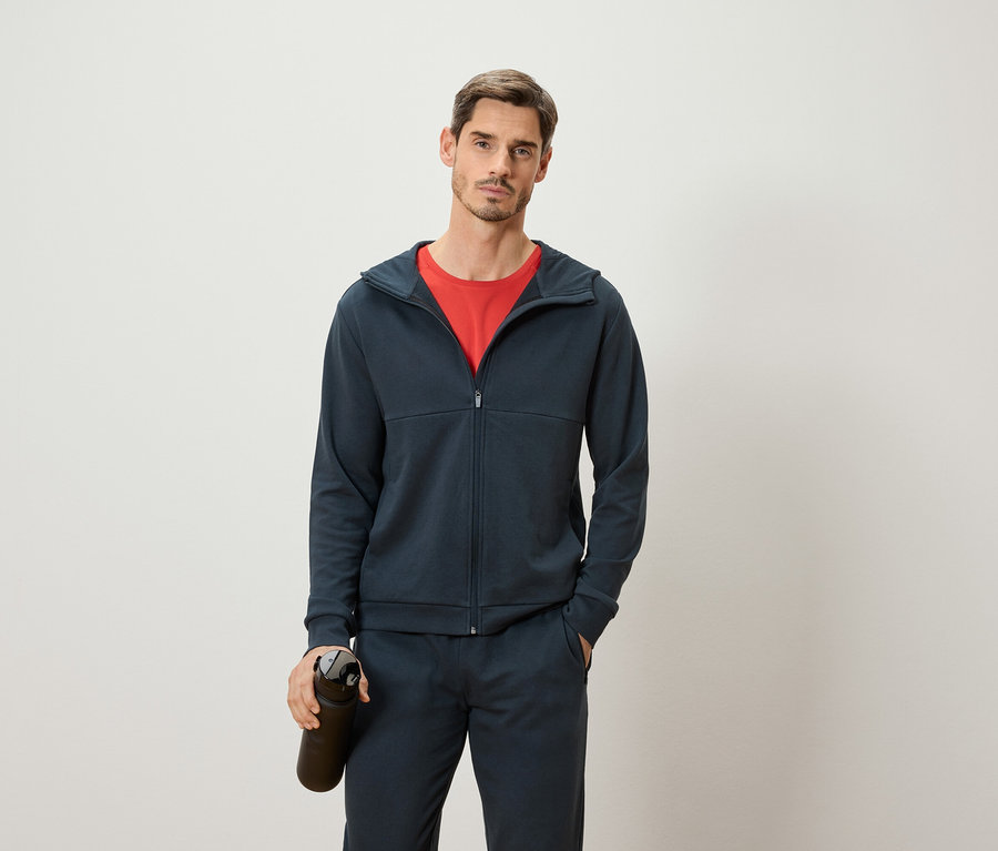 Un homme portant un t-shirt rouge, un sweat à capuche bleu marine et un pantalon de survêtement tient une bouteille d'eau sombre sur un fond blanc.