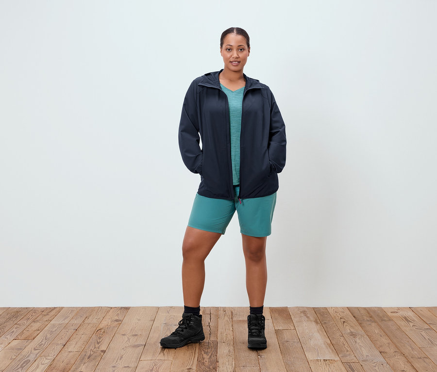 Une femme se tient debout, les mains dans les poches, devant un mur blanc. Elle porte une veste bleu marine, un t-shirt turquoise, un short turquoise et des bottes noires.