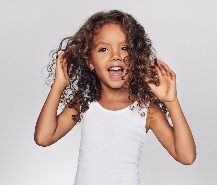 Une fillette aux cheveux bouclés et au débardeur blanc rit et touche ses cheveux devant un fond gris.