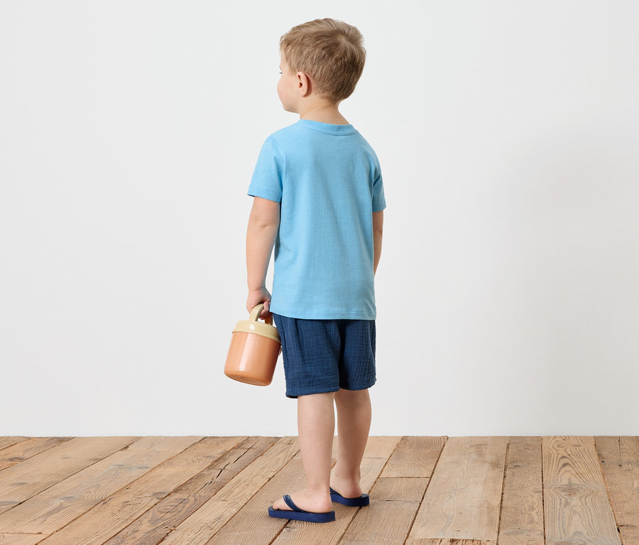 Un garçon vu de dos porte un t-shirt bleu, un short et des tongs bleues, et tient un récipient marron à la main.