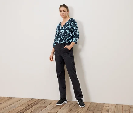 Femme debout avec la main dans la poche, portant un chemisier à motifs bleus et un pantalon noir.