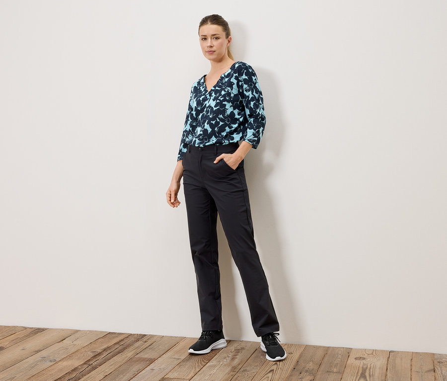 Femme debout avec la main dans la poche, portant un chemisier à motifs bleus et un pantalon noir.