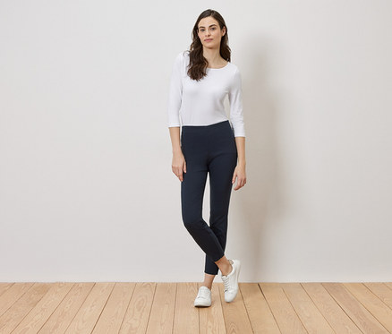 Une femme se tient devant un mur blanc, portant un haut blanc, un legging bleu marine et des baskets blanches.