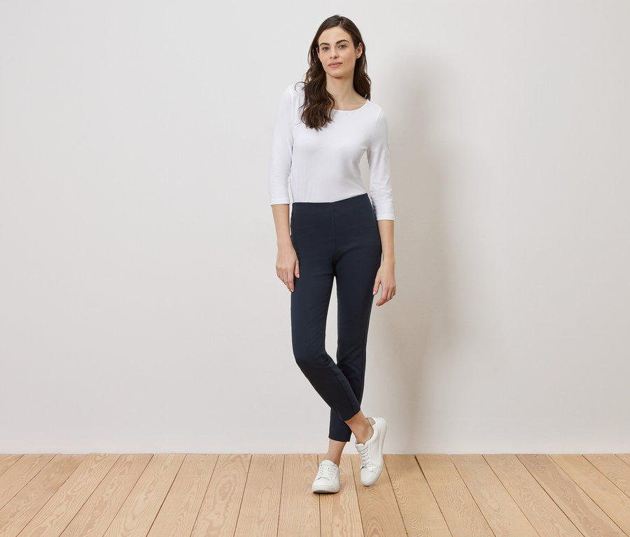 Une femme se tient devant un mur blanc, portant un haut blanc, un legging bleu marine et des baskets blanches.