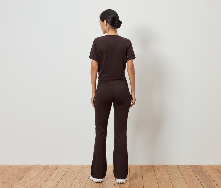 Une femme portant un chignon porte un t-shirt marron et un pantalon évasé, vue de dos sur un plancher en bois devant un mur blanc.