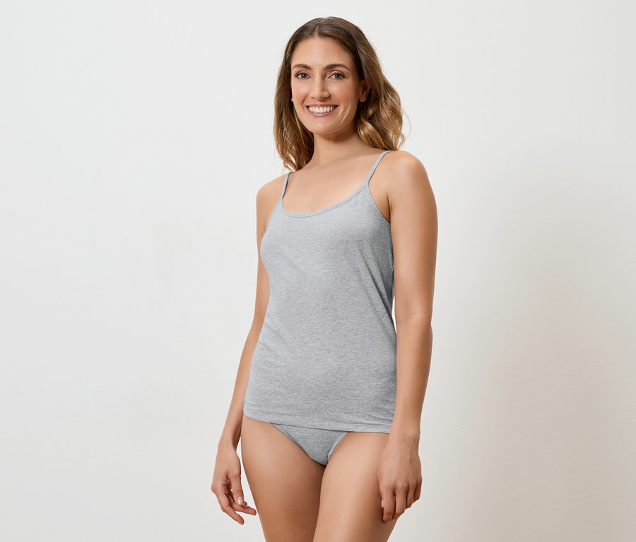 Une femme souriante aux longs cheveux bruns porte un débardeur et une culotte gris sur fond blanc.