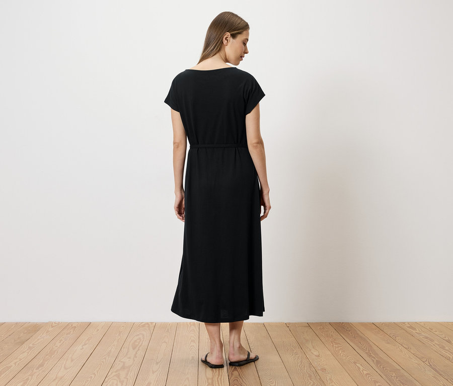 Une femme, vue de dos, porte une robe noire ceinturée arrivant aux chevilles et des sandales plates.