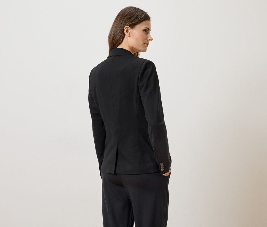 Femme vue de dos portant un blazer et un pantalon noirs, une main dans la poche, regardant par-dessus son épaule.