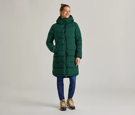 Une femme se tient sur un fond gris, portant un long manteau matelassé vert à capuche, un jean bleu et des bottes marron.