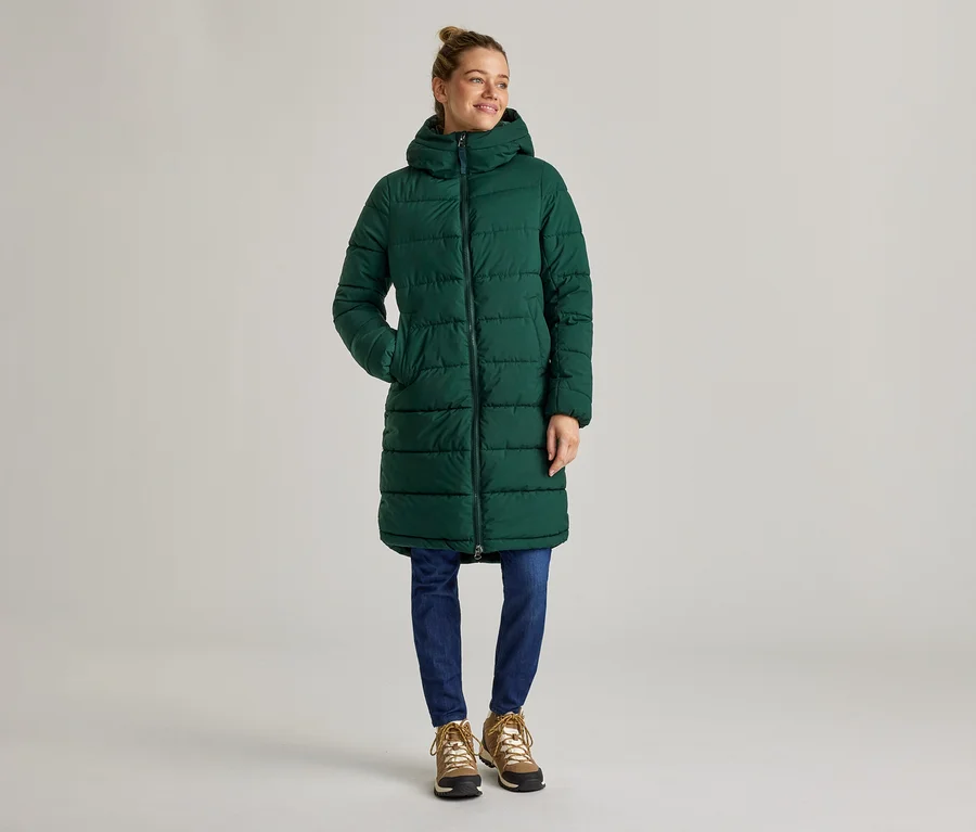 Une femme se tient sur un fond gris, portant un long manteau matelassé vert à capuche, un jean bleu et des bottes marron.