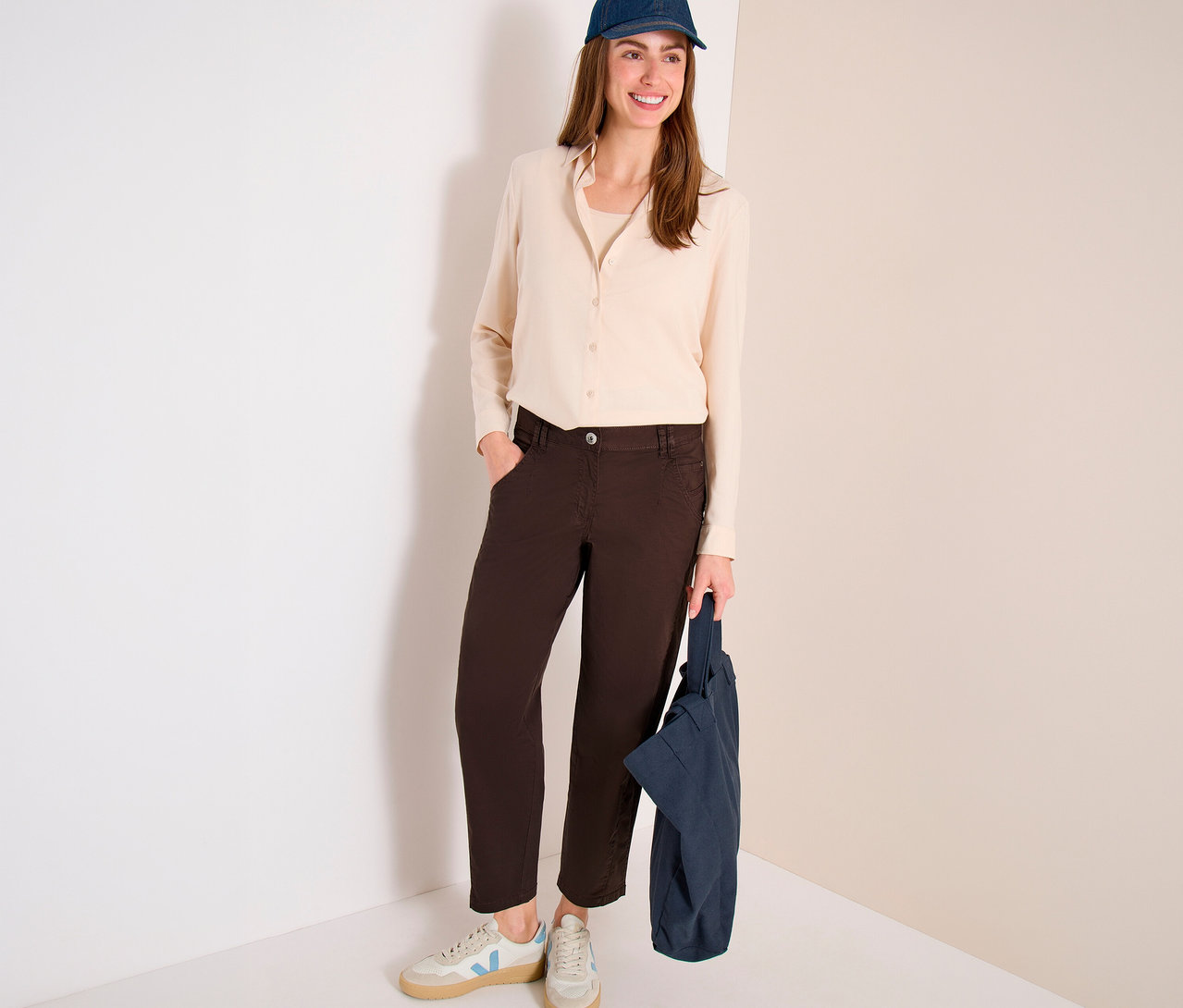 Une femme souriante est debout, vêtue d'une chemise claire, d'une casquette de baseball bleu foncé et d'un pantalon barrel CECIL « Scarlett » marron, tenant un sac fourre-tout bleu foncé.
