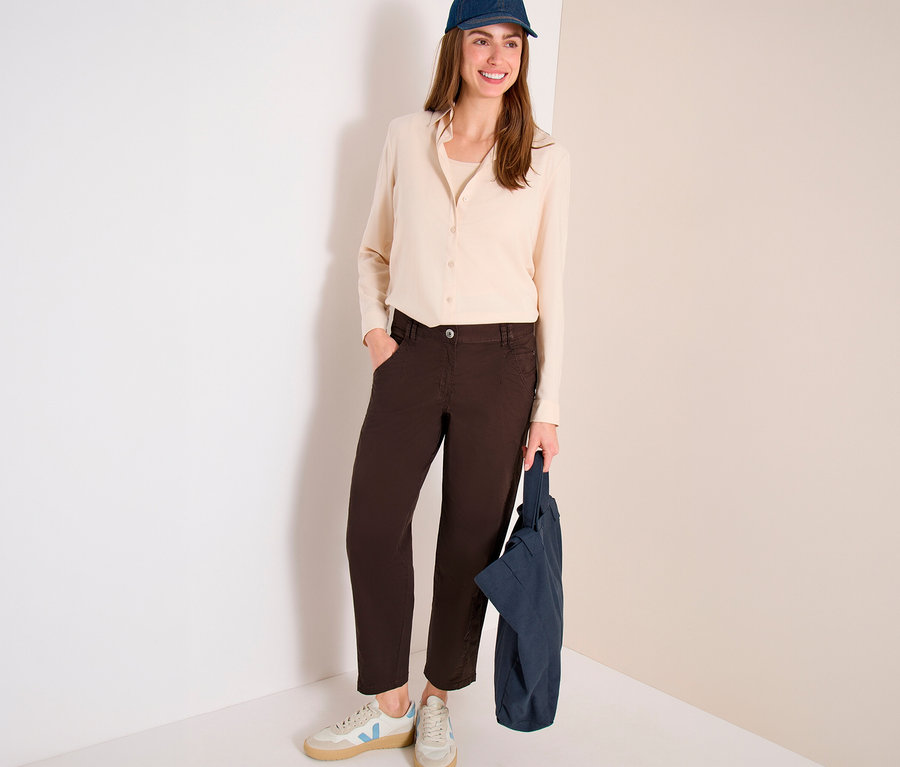 Une femme souriante est debout, vêtue d'une chemise claire, d'une casquette de baseball bleu foncé et d'un pantalon barrel CECIL « Scarlett » marron, tenant un sac fourre-tout bleu foncé.