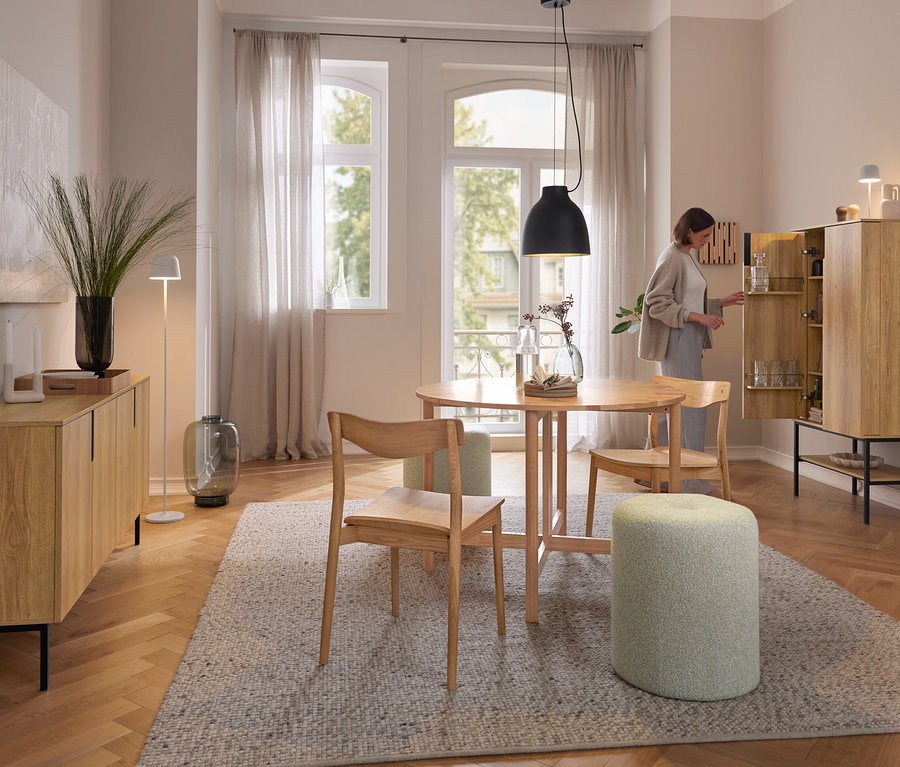 Une femme ouvre une armoire en bois clair dans une salle à manger. Au-dessus d'une table ronde en bois, une suspension noire est accrochée. Sur l'armoire, une lampe de table à LED rechargeable Eglo « Mannera » blanche.