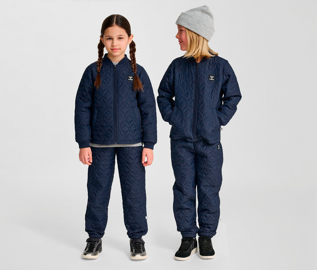 Deux enfants portent des ensembles hummel® « HMLSOBI Thermo-Set » bleus matelassés, composés d'une veste et d'un pantalon. Un enfant porte un bonnet gris.