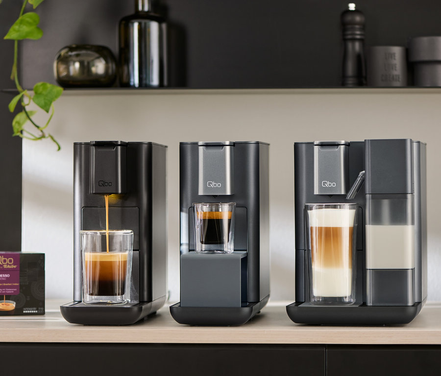 Trois machines à café Qbo sont posées sur un comptoir, dont les modèles Qbo MILK MASTER Gentle Grey, ICONIC Dark Chrome et ESSENTIAL Dark Stone. Préparation d'un expresso.