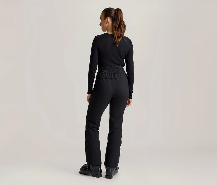 Femme vue de dos, portant un haut noir à manches longues, un pantalon de ski noir à taille élastique et des chaussures de ski noires. Ses cheveux bruns sont attachés en queue de cheval.